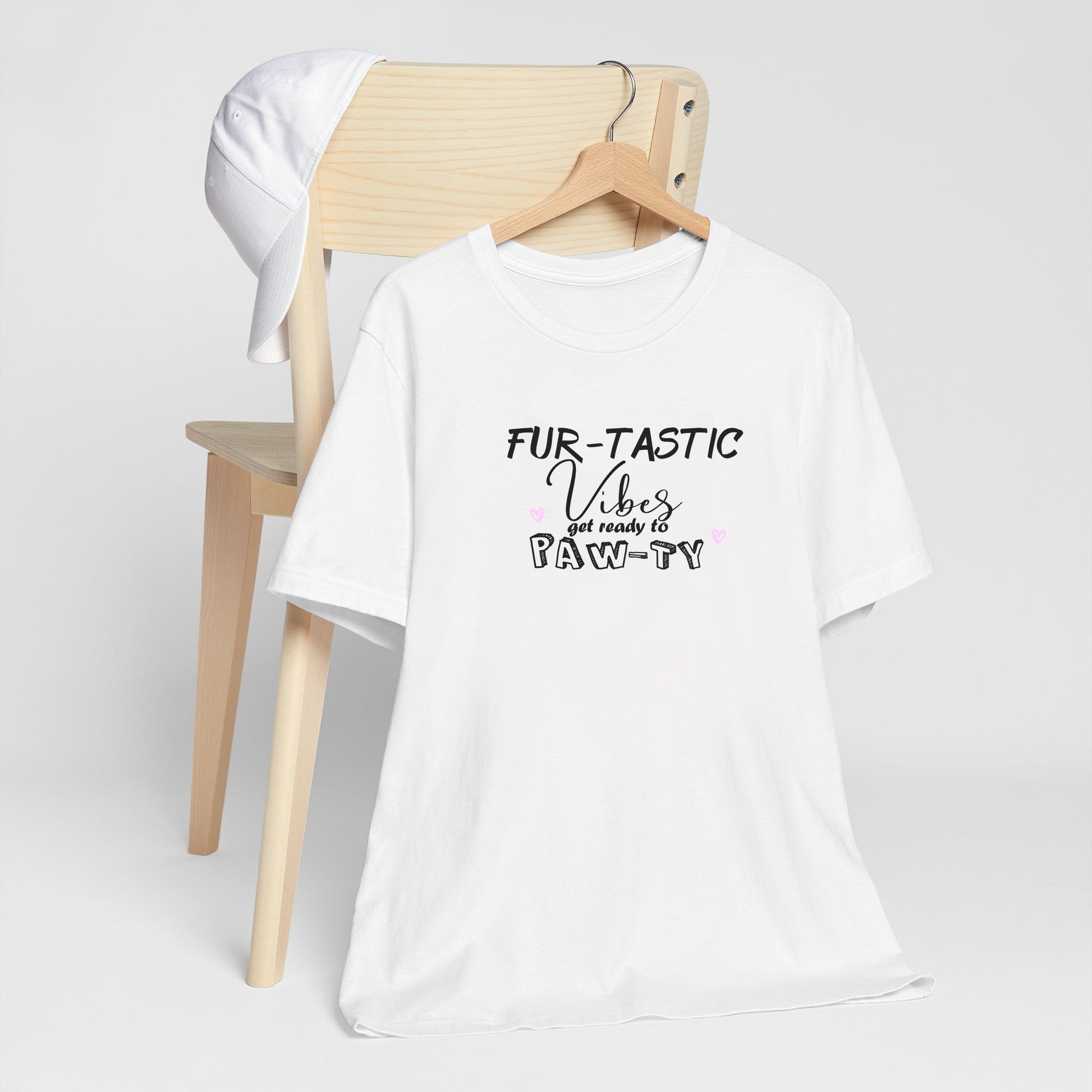 White crew neck short sleeved t-shirt, features a fun message "fur-tastic vibes get ready to paw-ty black graphic with cute pink hearts. T-shirt hanging on chair shown in close-up to highlight print and size