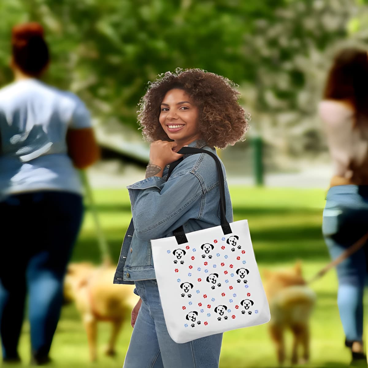 Woman standing in a dog park carry a on her shoulders a white spun canvas fabric tote bag, with black handle stripped, on the tote is a cute graphic cartoonist happy puppy face. surrounded by colorful paws