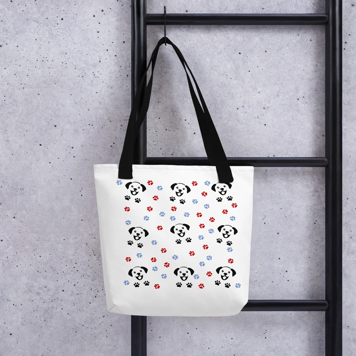 Tote bag hanging on a hook. Black handle and a white spun canvas fabric tote bag, with black handle stripped, on the tote is a cute graphic cartoonist happy puppy face, surrounded by colorful paws.