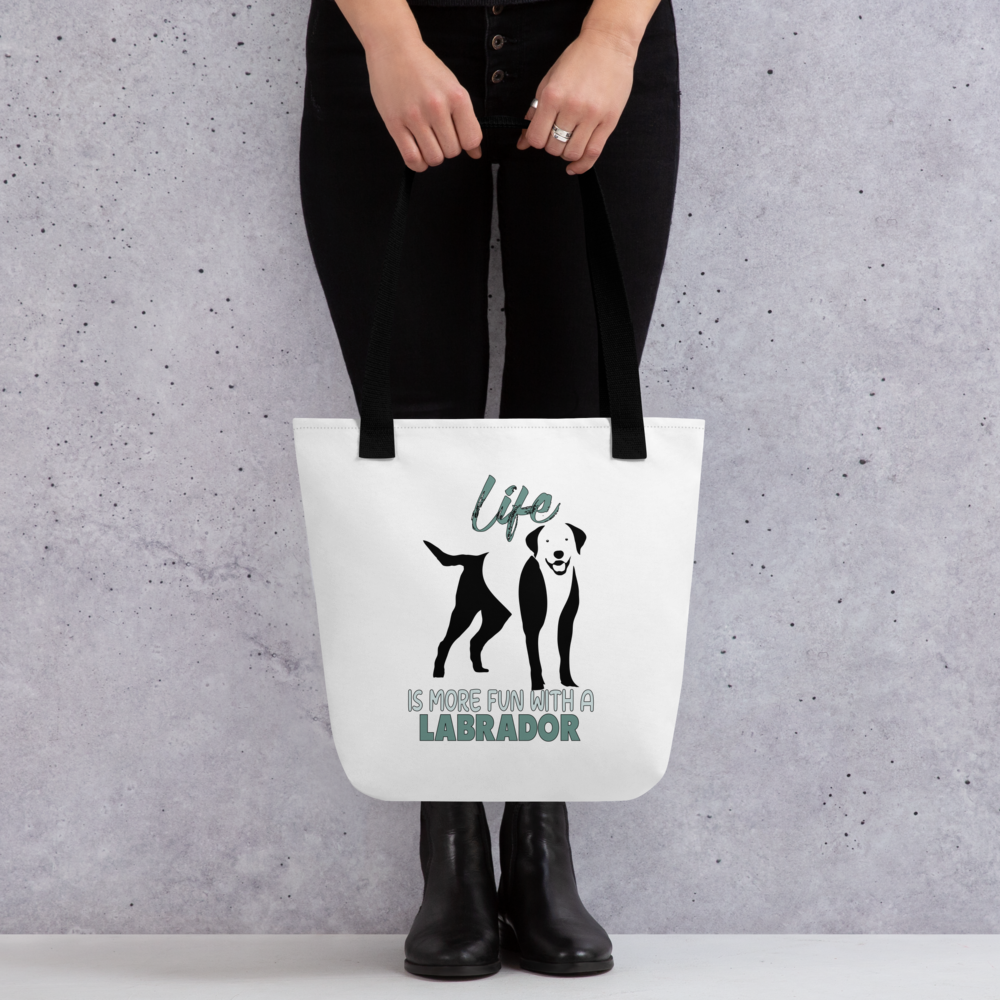 White spun canvas fabric tote bag , on the bag is a black one-line graphic print silhouette of a cute Labrador dog standing with its tongue out. Above the dog is the text " LIFE" in cap and underneath the dog is the text " is more fun with a Labrador " in cap" 