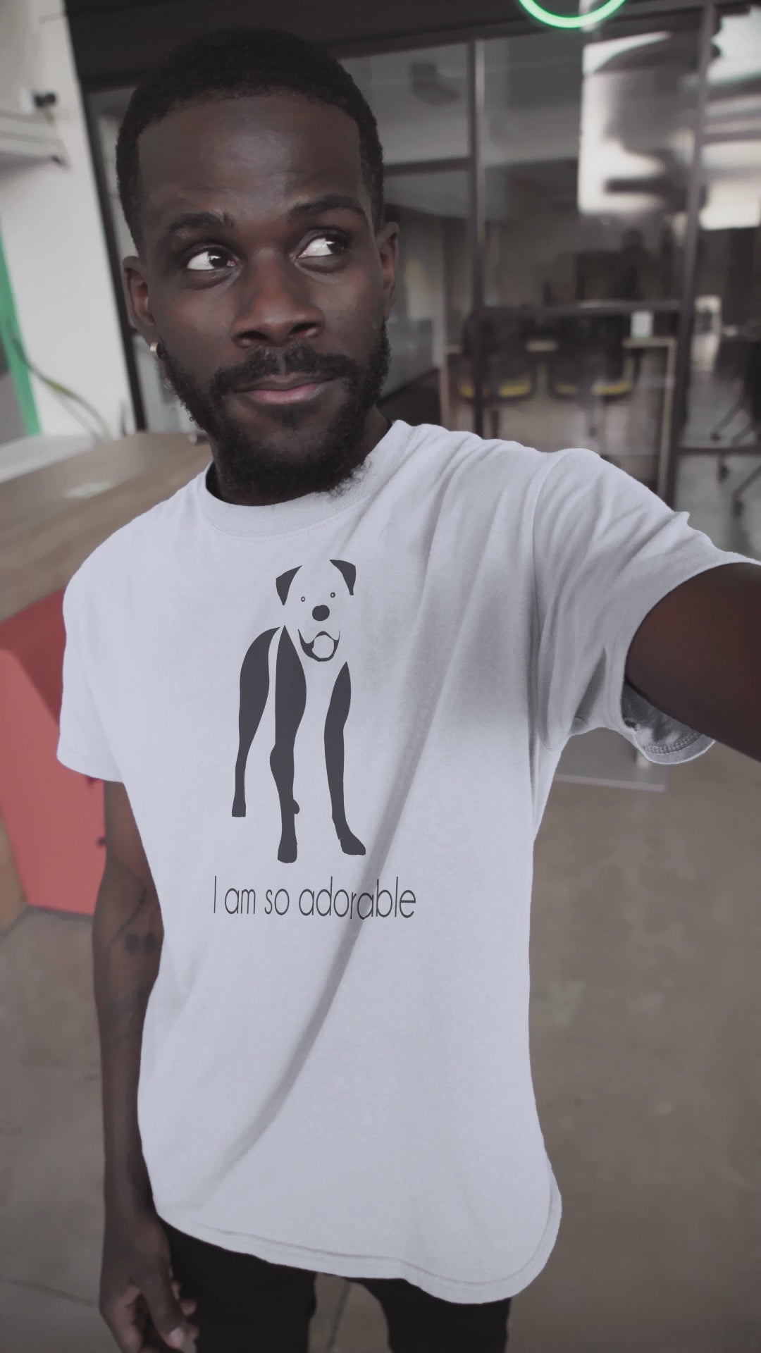 close up video of a smiling man with beard, wearing a white crew neck short sleeve t-shirt, with a gaphic silhouette print, feature a one-line black and white graphic of a boxer dog standing with it tounge our, below the dog is the text " i am so adorable" in black text style.