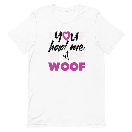 Unisex short sleeve crew neck crisp white color t-shirt, feature a classic quote you had me at woof, the letter o, is a pink colred heart with dog paw, there is underlay of thentext woof, repeated several times.