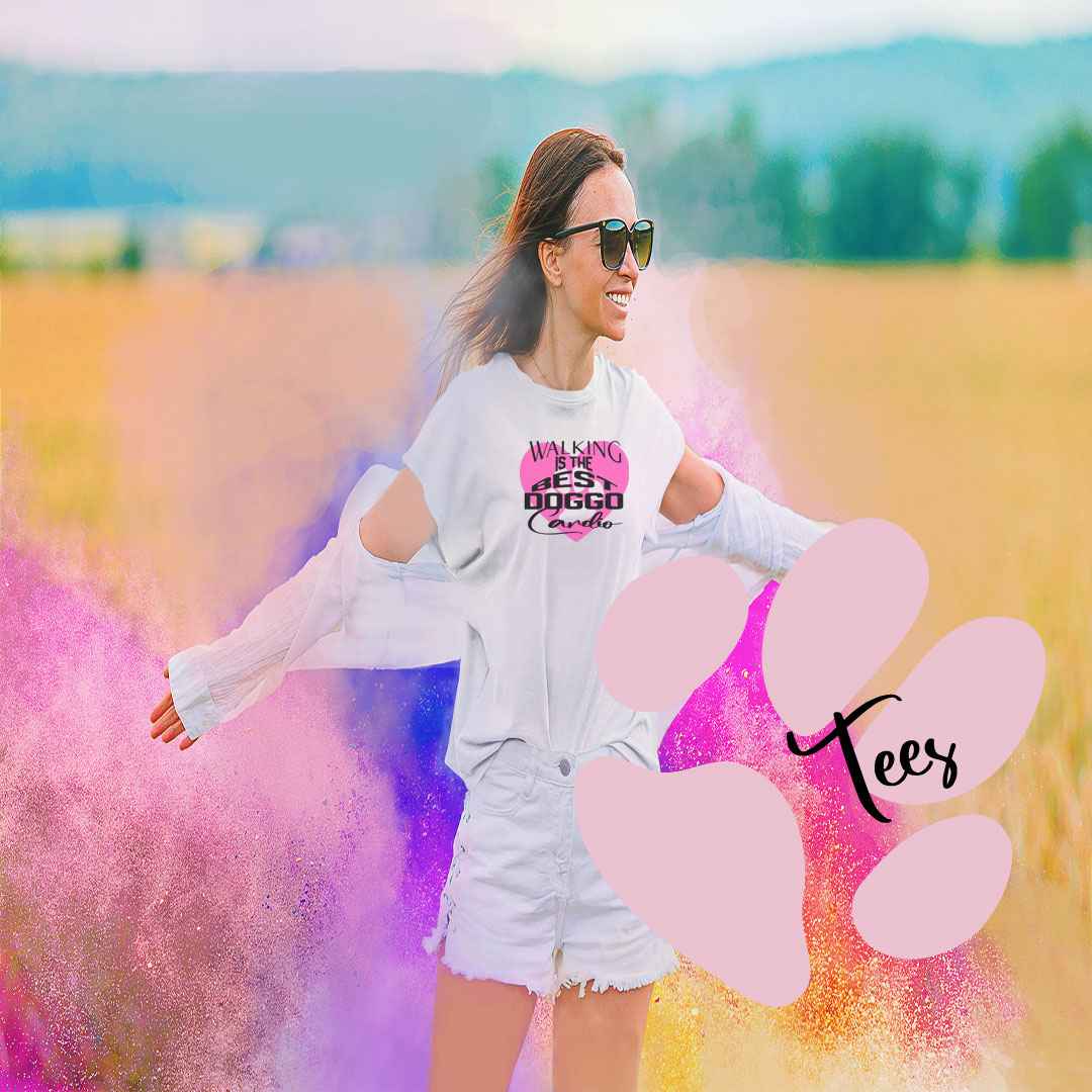 Close up of women carring a graphic white short sleeve tee with purple shaped heart and dog paws, on top of the heart is the text"walking is the best doggo cardio"stay" The women is outside on a farm field, spreading her arms in the air, happy face and dark sun glasses. The photo is square, background blurred in different bright light colours, in the right side corner is a large pink dog paw  topped by the word "Tees"