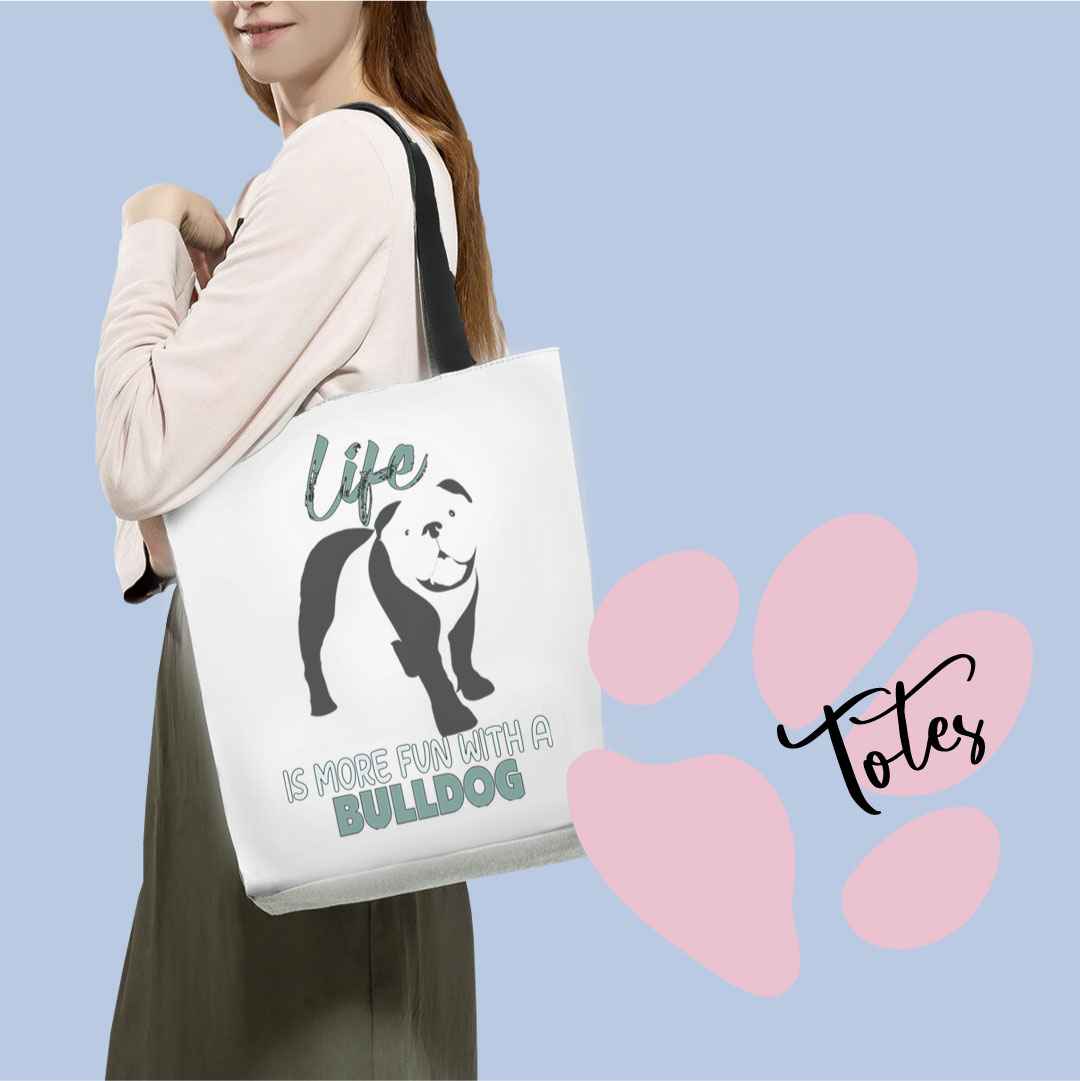 Close up of women carring a graphic tote bag with black striped handle, the graphic shows a cute bulldog standing, above the dog is the text "life in cap, and below is the text "is more fun withn a Bulldog" The photo is square, back ground bluish, with a large pink dog paw in the corner, top by the word "totes"