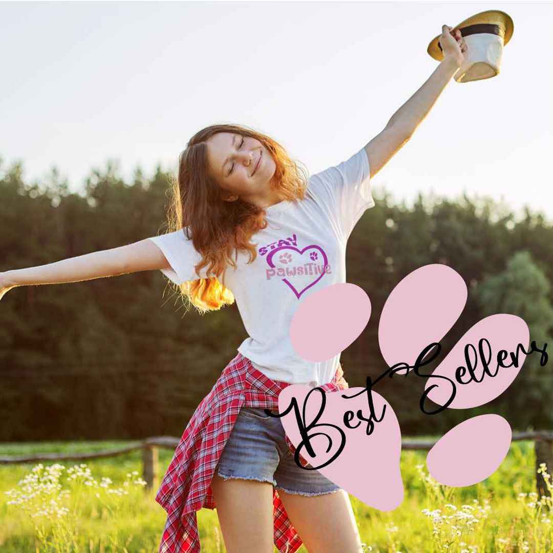 Close up of women carring a graphic white short sleeve tee with purple shaped heart and dog paws, above is the text"stay" and in the middle of the heart is the text "pawsitive" The women is outside on a farm field, spreading her arms in the air, happy face. The photo is square, background blurred, with a large pink dog paw in the corner, top by the word "best Sellers"