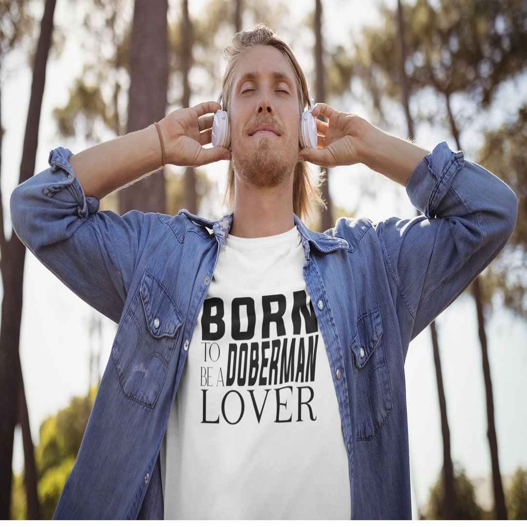 White cotton crew neck unisex shirt, short sleeve, on the apparel is a black and white graphic print with the text BORN TO BE A DOBERMAN LOVER in capital letters in different text styles