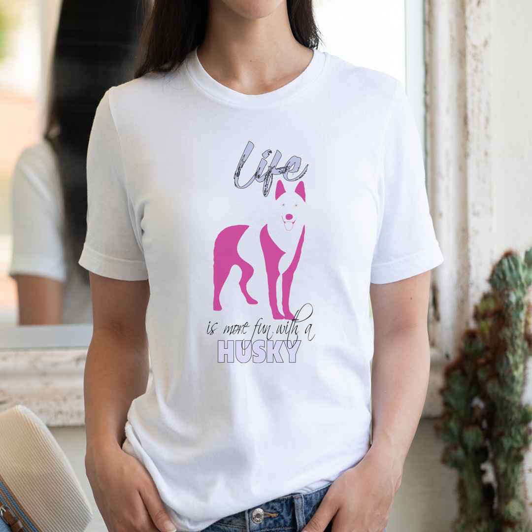 White cotton crew neck unisex  short sleeve tshirt, on the apparel is a graphic print silhouette of a cute adorable SIBERIAN Husky standing , the dog silhouette is in pink color, above the dog is the text " Life" written in tilted purple graphic letter, below is the text " is more fun with a Husky" in capital letter, the text "Husky" stands out with its pink color