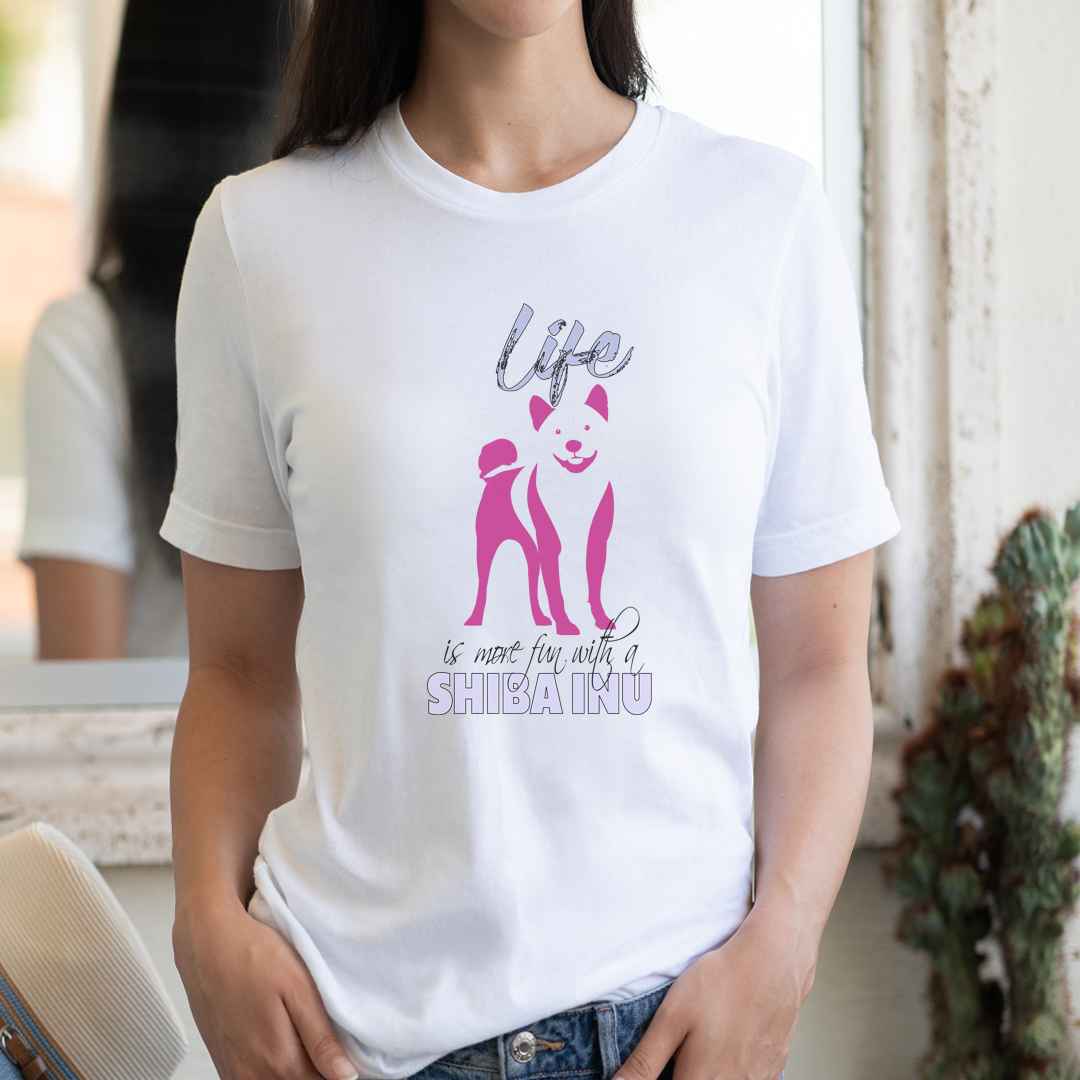 White cotton crew neck unisex  short sleeve tshirt, on the apparel is a graphic print silhouette of a cute adorable Shiba standing , the dog silhouette is in pink color, above the dog is the text " Life" written in tilted purple graphic letter, below is the text " is more fun with a Shiba" in capital letter, the text "Shiba" stands out with its pink color