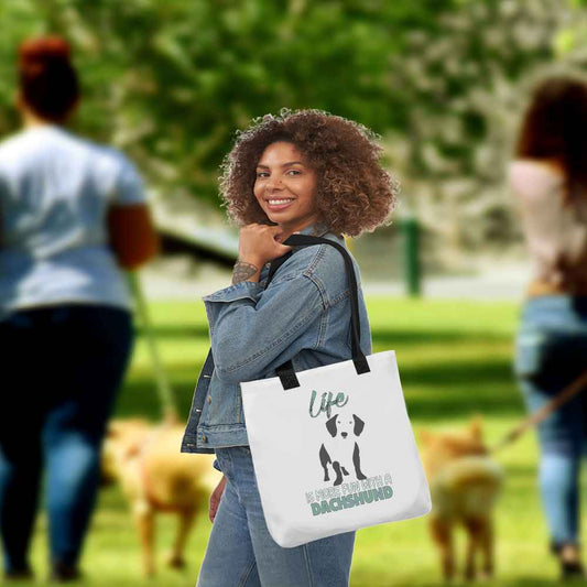 Young women walking in  a park, wearing on her shoulder a white tote polyester canvas bag, with black stripped handle, feature a black one-line Dachshund print with life quote ”life is more fun with a Dachshund”. Printed front and back.