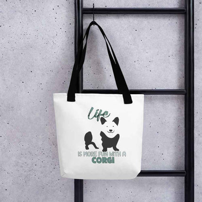 White spun canvas fabric tote bag , on the bag is a black one-line graphic print silhouette of a cute Welsh Corgi dog standing with its tongue out. Above the dog is the text " LIFE" in cap and underneath the dog is the text " is more fun with a Corgi " in cap" 